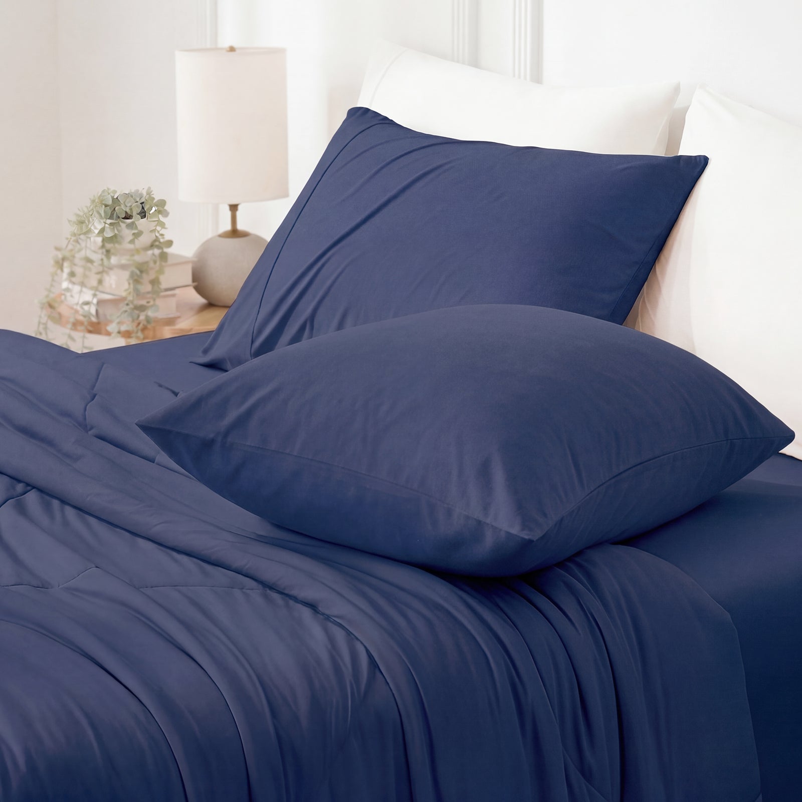 Dark blue bedding set on a bed with pillows and a lamp in the background.#color_Dark Blue